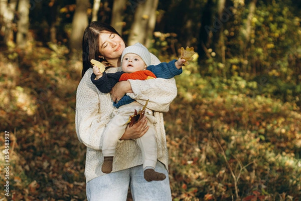 Obraz Happy mother hugging cute baby son in autumn woods. Motherhood. Stylish woman with adorable child in cozy warm sweaters embracing and relaxing in fall park