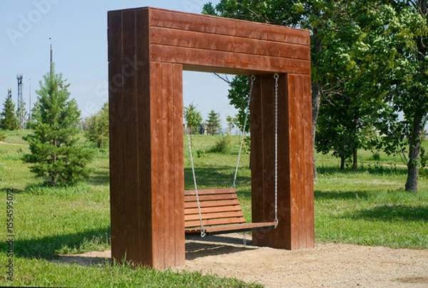 Obraz A wooden swing bench suspended by chains within a modern park installation surrounded by lush greenery. Photo