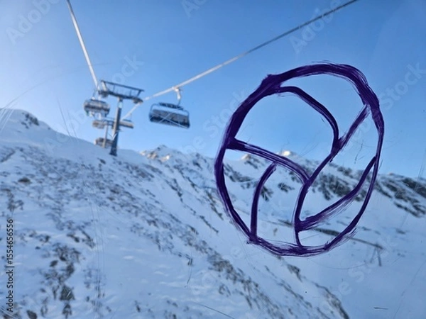 Fototapeta A ski lift is in the background of a snowy mountain. A purple volleyball is drawn on a window on ski resort