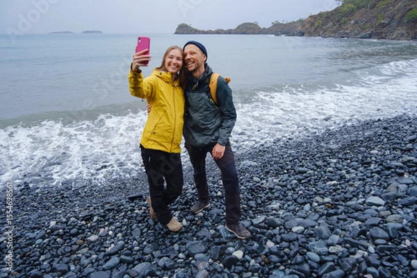 Fototapeta Couple enjoys Turkish coastline and taking selfie on beach in Winter. walking on stones, watching sea waves, relaxing together, slow romantic moment near calm blue water under soft winter light.