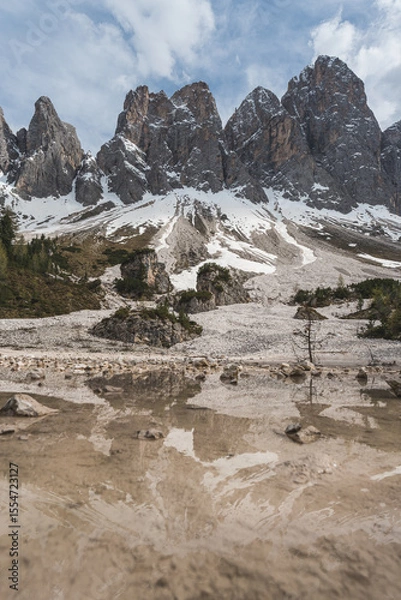 Fototapeta Spring snowmelt and mountain reflection at foot of Geisler peaks in the Dolomites, with cloudy sky and alpine forest landscape.