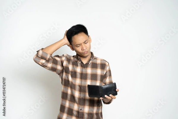 Fototapeta A man wearing a plaid shirt, expressing concern as he looks at his empty wallet, conveying financial strain.