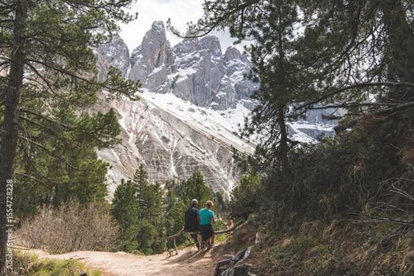 Fototapeta Couple sitting on bench in alpine forest enjoying snowy Dolomites mountain view in Italy.