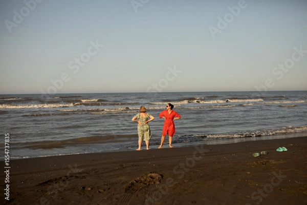 Fototapeta Two women enjoying a joyful seaside moment during vacation, barefoot on the beach, sharing laughter and sunshine. A memory of freedom, travel, and connection