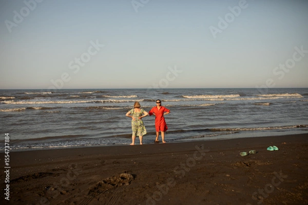 Fototapeta Senior mother and adult daughter enjoy quality time by the sea, barefoot on a sunny beach, sharing smiles and the joy of travel, connection, and warm memories.