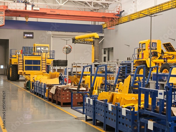 Fototapeta A factory with a lot of yellow and blue machinery trucks. The machinery is being built and the workers are busy