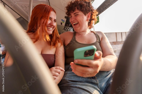 Fototapeta Two women riding public transit, smiling together while scrolling smartphones, sharing casual camaraderie during urban commute