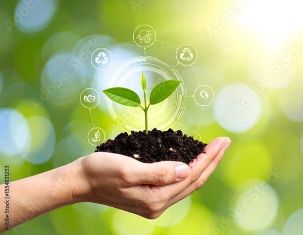 Fototapeta A hand holds a small plant sprouting from rich soil, with eco-icons like recycling and energy symbols floating above. The blurred green-yellow background adds a hopeful, environmental tone.