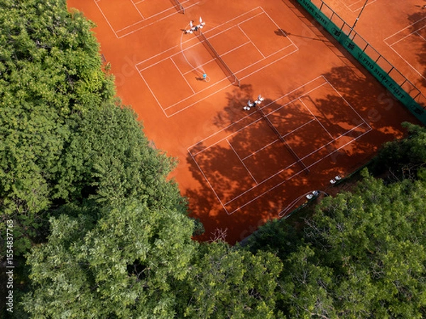 Fototapeta Aerial view of red clay tennis courts surrounded by lush green trees. Empty courts with shadows and equipment create a peaceful and organized sports atmosphere