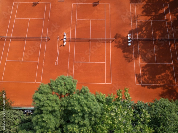 Fototapeta Aerial view of red clay tennis courts surrounded by lush green trees. Empty courts with shadows and equipment create a peaceful and organized sports atmosphere