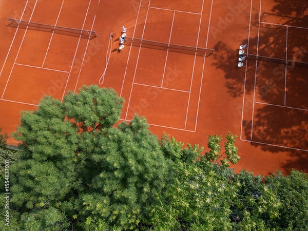 Fototapeta Aerial view of red clay tennis courts surrounded by lush green trees. Empty courts with shadows and equipment create a peaceful and organized sports atmosphere