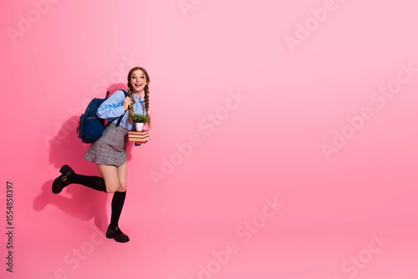 Fototapeta Happy female student holding a plant and boo book s while posing cheerfully against a vibrant pink background