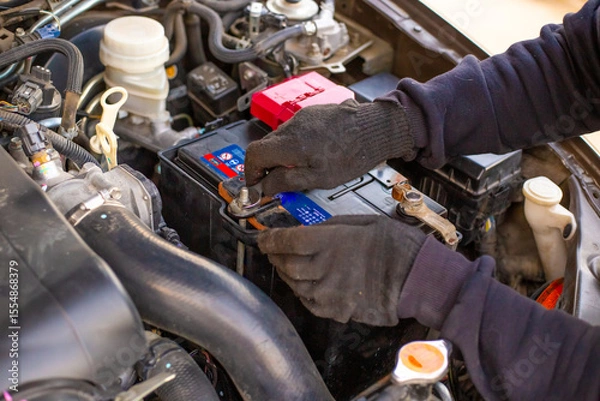 Fototapeta A man wearing gloves securely fastens a car battery terminal after installing it under the hood. Replacing a weak battery