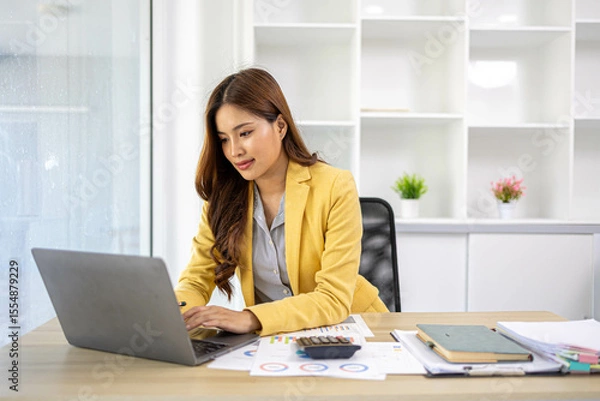 Fototapeta A businesswoman is working in her office. She is checking documents at her desk with a calculator and a mobile phone. She is looking at the calculator and thinking.

