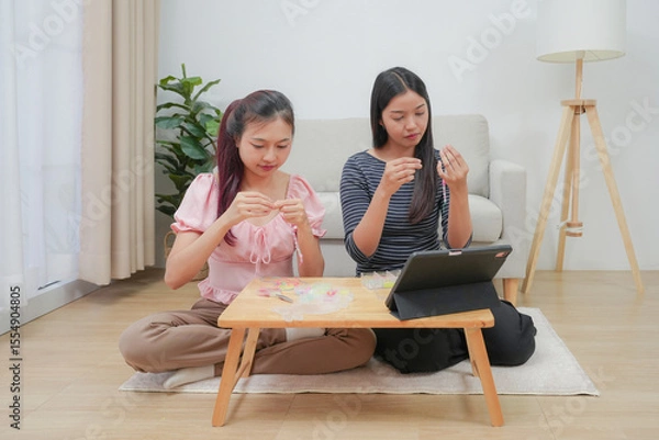 Fototapeta Two young asian women stringing beads on thread and choosing colorful beads from box to making handmade bracelet while spending time to creating jewelry handicraft and doing hobby lifestyle at home. 