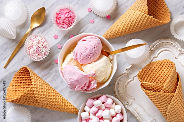 Fototapeta Ice cream ingredients view from above on a marble background. Ice cream in a bowl, ice cream cones, spoon, marshmallows, cupcake papers. Top view.