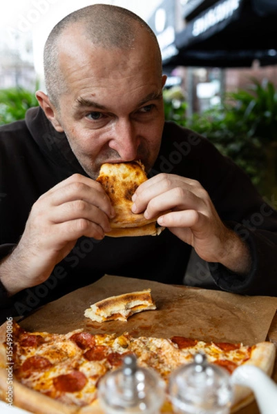 Fototapeta A man with a glass of dark beer in his hand enjoys a slice of pizza. There is a salt and pepper shaker and a kettle on the table.
