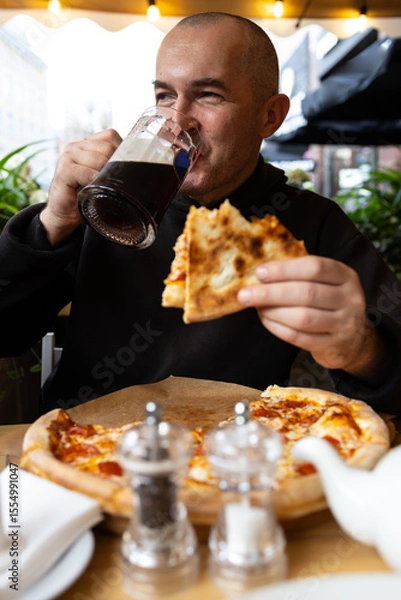 Fototapeta A man with a glass of dark beer in his hand enjoys a slice of pizza. There is a salt and pepper shaker and a kettle on the table.