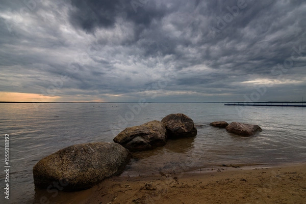 Obraz summer evening lake landscape. large coastal stones in calm water near sandy shore under cloudy dramatic sky with glow