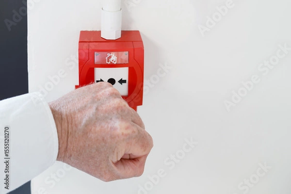 Fototapeta Office worker seen attempting to break the thin glass to activate a generic fire and smoke alarm seen within a commercial building. An emergency exit door is on the left of the image.