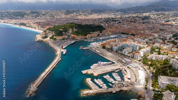 Fototapeta Wide aerial view of the Port of Nice, France, showing the coastline, marina with yachts, city buildings, and surrounding hills on a partly cloudy day.