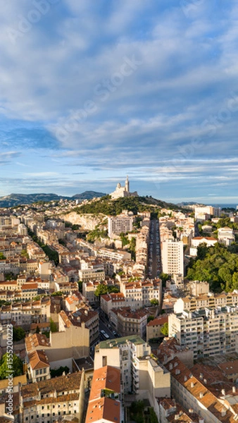 Fototapeta Drone aerial view of the city of Marseille, France, with the iconic Notre-Dame de la Garde basilica on a hilltop surrounded by urban buildings under a partly cloudy sky.