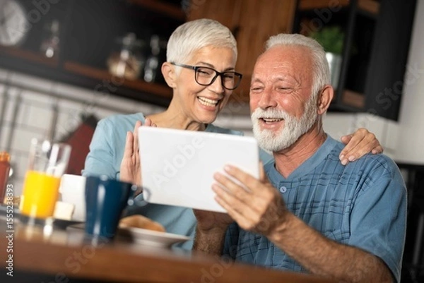 Obraz Happy older mature couple having breakfast using digital tablet in kitchen.