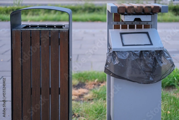 Fototapeta Close-up view of two different styles of public trash bins positioned on a sidewalk in a green urban environment.