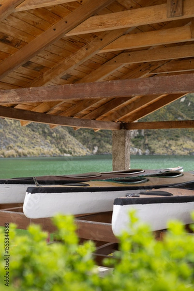 Fototapeta Tucked beneath a rustic roof, a group of kayaks rests silently by the lake. The scene speaks of calm moments before sunrise, still waters, and stories yet to be paddled.

