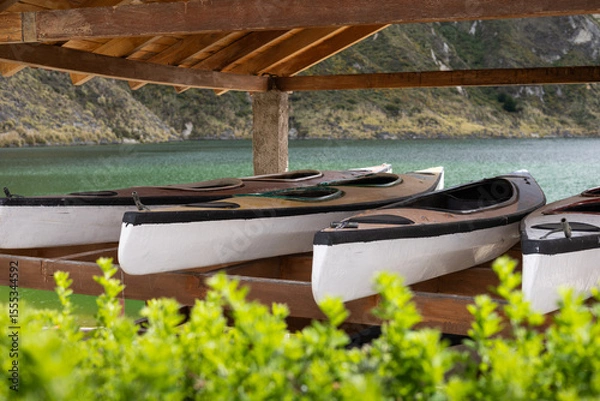 Fototapeta A line of kayaks rests under a wooden shelter by the lake, surrounded by mountains. The image captures a perfect pause before outdoor adventure in a stunning natural setting.

