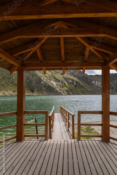 Fototapeta A captivating view from inside a wooden pier reveals intricate timber construction alongside a breathtaking panorama of the calm lake and lush mountains beyond.


