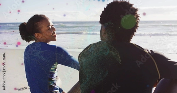 Obraz Sitting wetsuit-clad adult couple watching gentle ocean waves lapping shore at beach, sunny sky