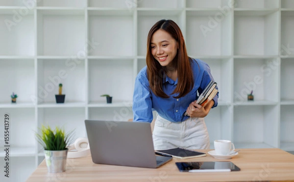 Obraz Smiling young asian woman sitting at desk working on laptop taking notes in notebook, happy millennial female studying online, watching webinar using computer and writing check list
