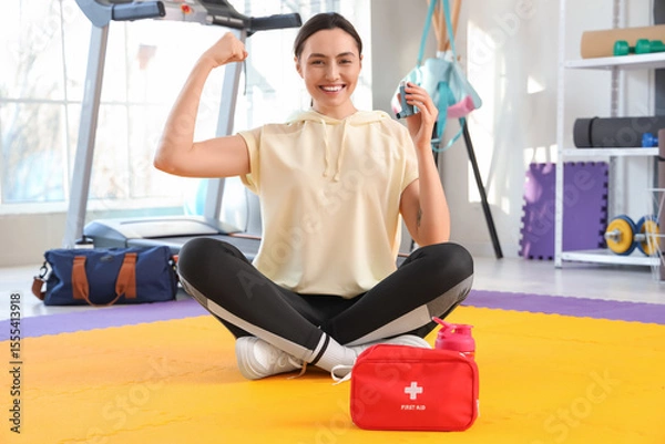Fototapeta Sporty young woman with inhaler and first aid kit showing muscles in gym