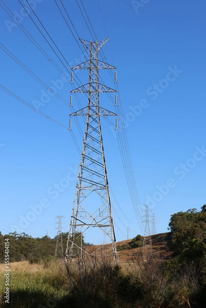 Fototapeta Transmission electricity tower in a grassy landscape with trees under a clear blue sky