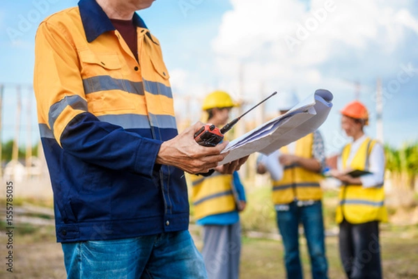Fototapeta At a table on a construction site, an elderly male safety expert, a middle-aged man, and two young Asian women gather around a laptop to actively document, coordinate plans, and execute projects