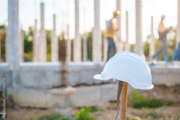Fototapeta An Asian middle-aged male engineer wearing a yellow safety helmet and reflective vest uses a walkie-talkie at an active construction site, coordinating tasks amid steel beams and building framework