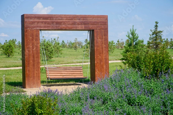 Obraz A wooden bench swing hanging by metal chains from a large timber frame in a sunny urban park, surrounded by green grass, purple blossoms, and young trees. Photo