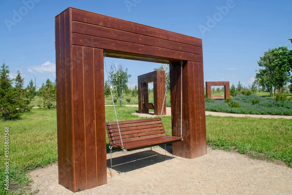 Obraz A large wooden swing bench suspended by metal chains from a sturdy timber frame in a green park, with additional swing installations receding into the background. Photo