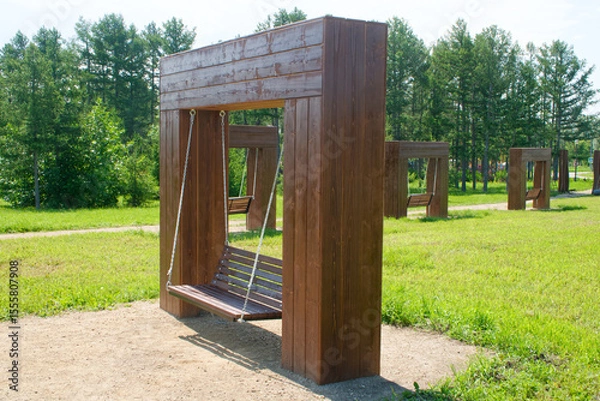 Obraz A series of wooden swing benches suspended by metal chains from large timber frames in a sunlit urban park, surrounded by grass and trees. Photo