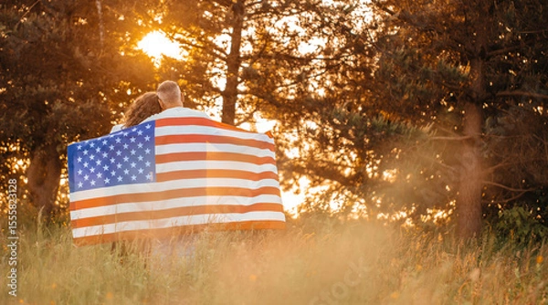 Obraz A couple embraces peacefully in a sunlit meadow wrapped in an American flag, surrounded by tall grass and glowing trees in the warm golden light of a summer sunset