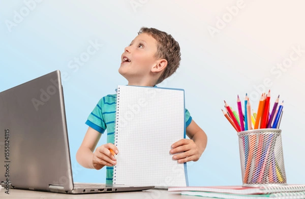 Obraz A cheerful young boy shows his notebook during an online learning session, sitting at a desk with a laptop and colorful pencils, ready for a fun and creative school day