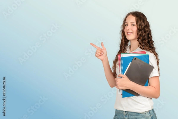 Obraz A confident teenage girl with long curly hair points to the side while holding school notebooks and a tablet, ready for a successful and productive academic day