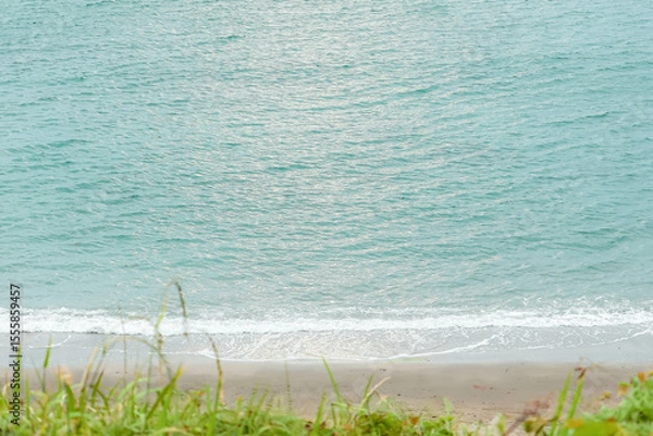 Obraz エメラルドグリーンの海を引き立てる緑色の草の間から、崖下のビーチを眺めた風景