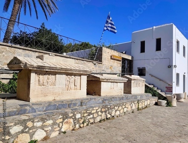 Fototapeta PAROS, GREECE - JUNE 28TH 2025: Exterior view of the Archaeological Museum of Paros in Parikia. It features indoor and outdoor exhibits of ancient artifacts, statues, and sarcophagi.