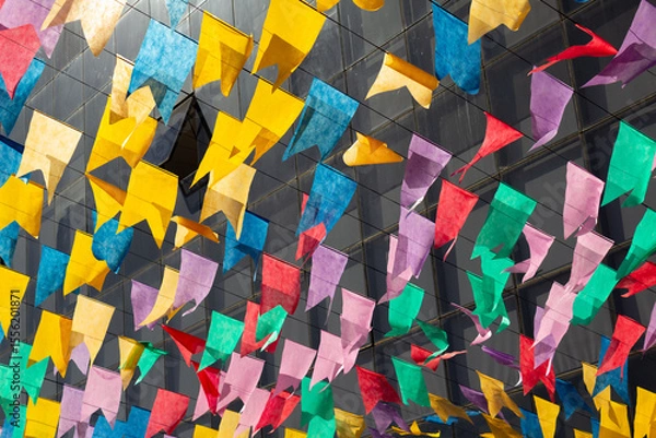 Fototapeta Bandeirolas coloridas, penduradas ao ar livre dançando ao vento com janelas de um predio desfocado ao fundo.