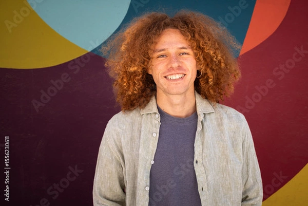 Fototapeta Portrait of smiling young man with curly hair standing in front of vibrant colorful mural. Urban style, relaxed expression, casual outfit, youth culture, creativity and artistic background.
