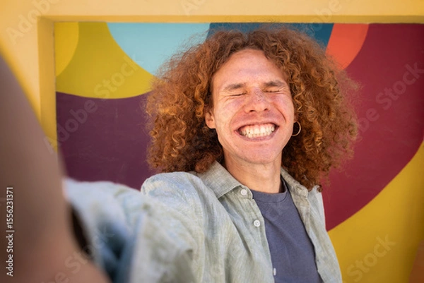 Fototapeta Cheerful student with curly hair taking a joyful selfie with eyes closed and big smile in front of colorful mural. Urban youth, university lifestyle, fun expression and digital personality.