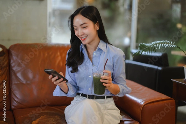 Fototapeta Young asian businesswoman using mobile phone and drinking healthy green vegetable smoothie while sitting on sofa in cafe