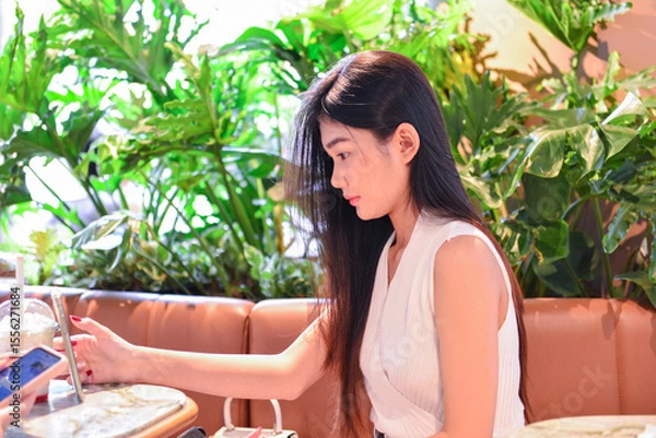 Obraz Asian Woman Using Smartphone in a Modern Café Surrounded by Plants